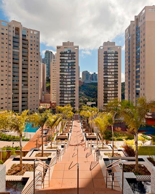Vista aérea de condomínio residencial com escadaria central, palmeiras, espelhos d'água, piscina e torres de apartamentos em dia ensolarado